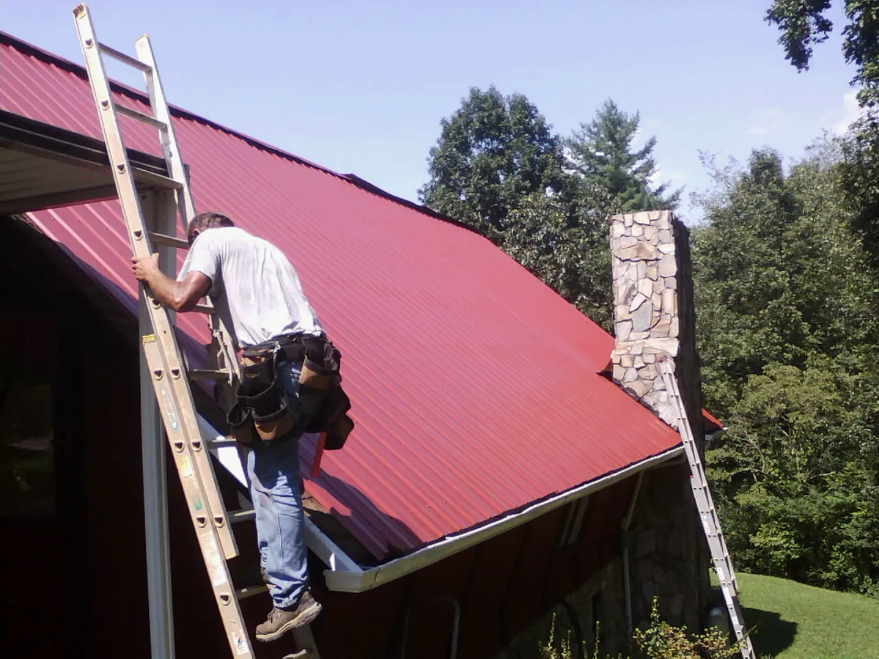 Roofer on a ladder beside a red metal roof and stone chimney — Soffit & Fascia Repair by Roofing Brothers in Woodrow